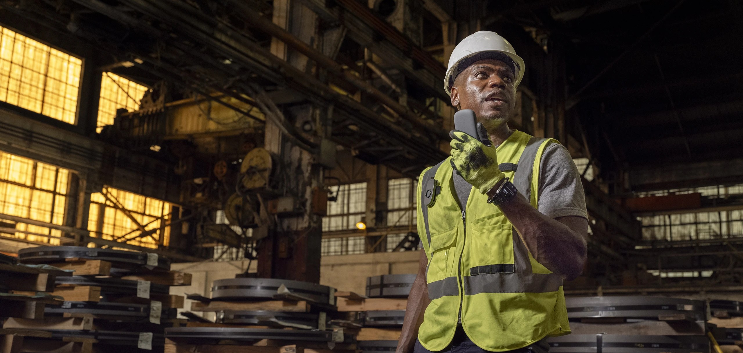 A frontline worker in a hard hat and hi-vis vest speaking into a Relay radio inside an industrial facility